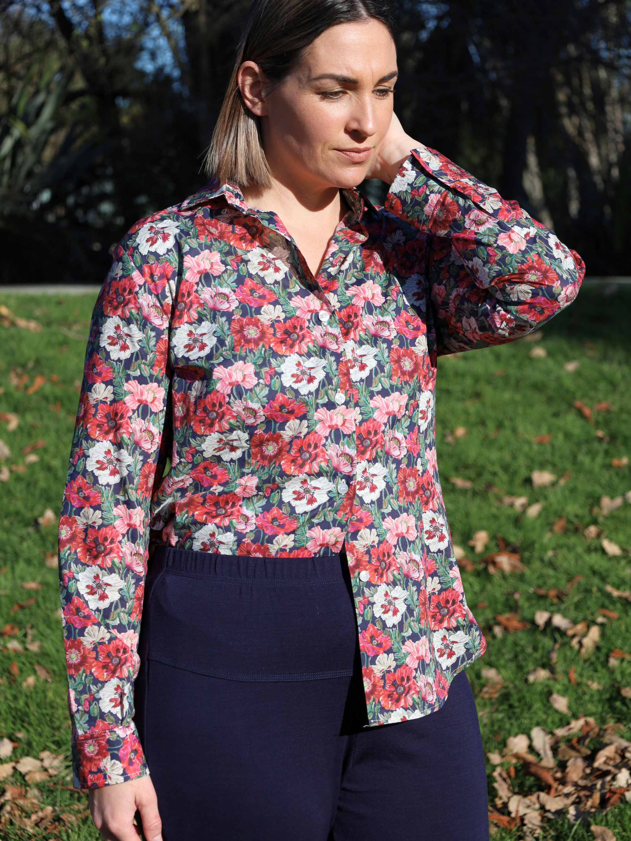 Woman wearing a floral blouse standing outdoors on a grassy area with trees in the background.