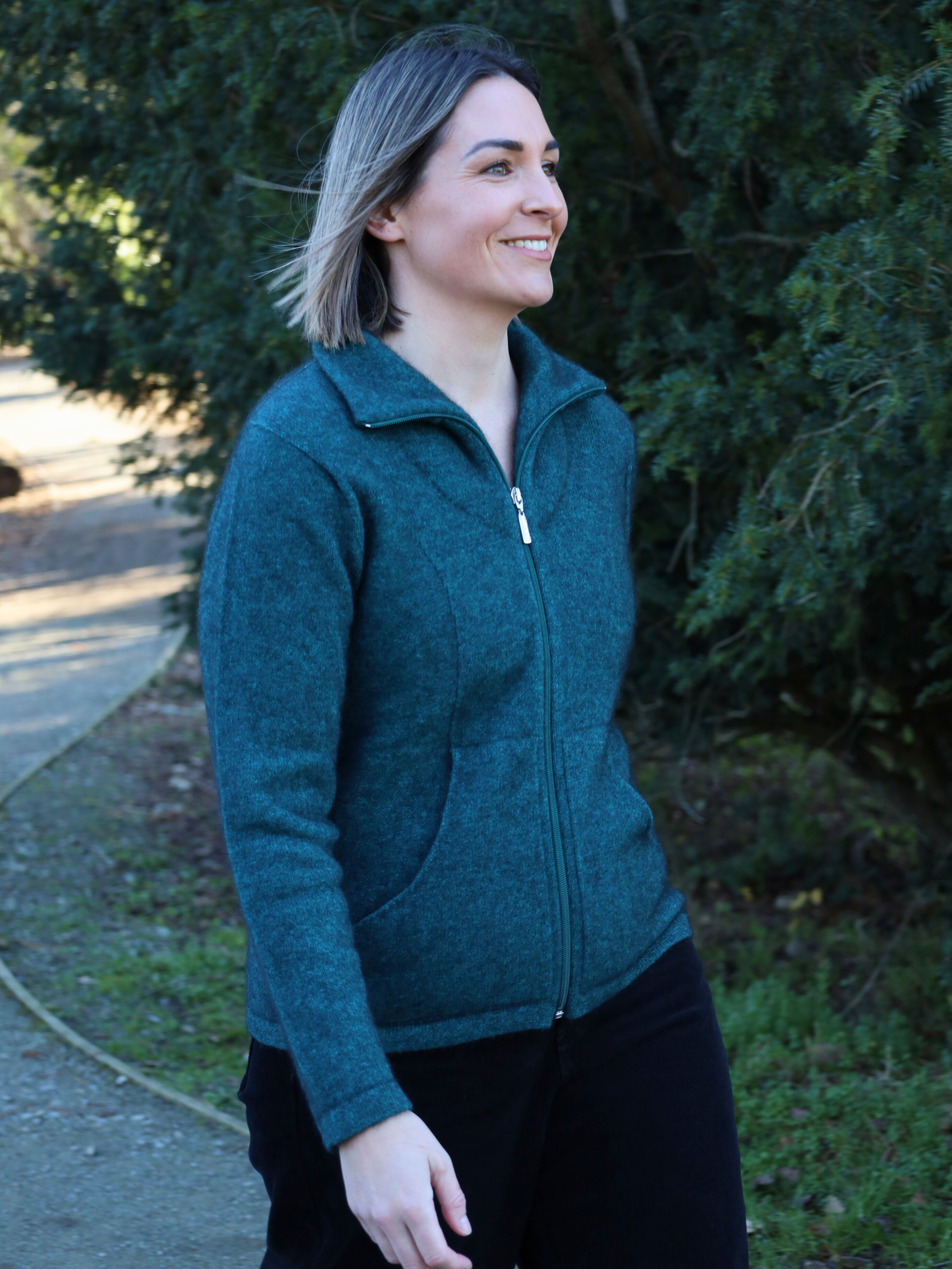 Woman wearing a teal jacket standing outdoors with greenery in the background